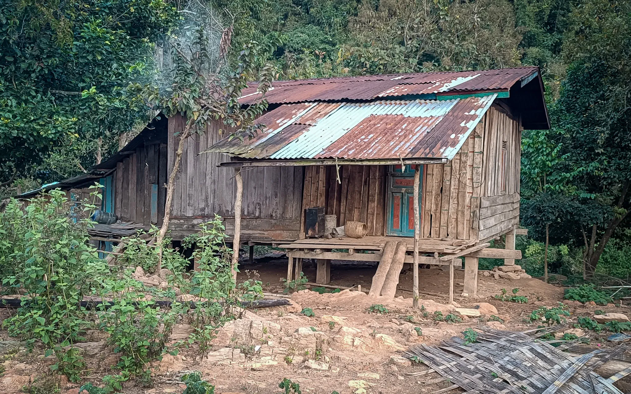 An abandoned wooden stilted house with a rusted tin roof surrounded by overgrown vegetation at a deserted settlement on the Nam Ou river