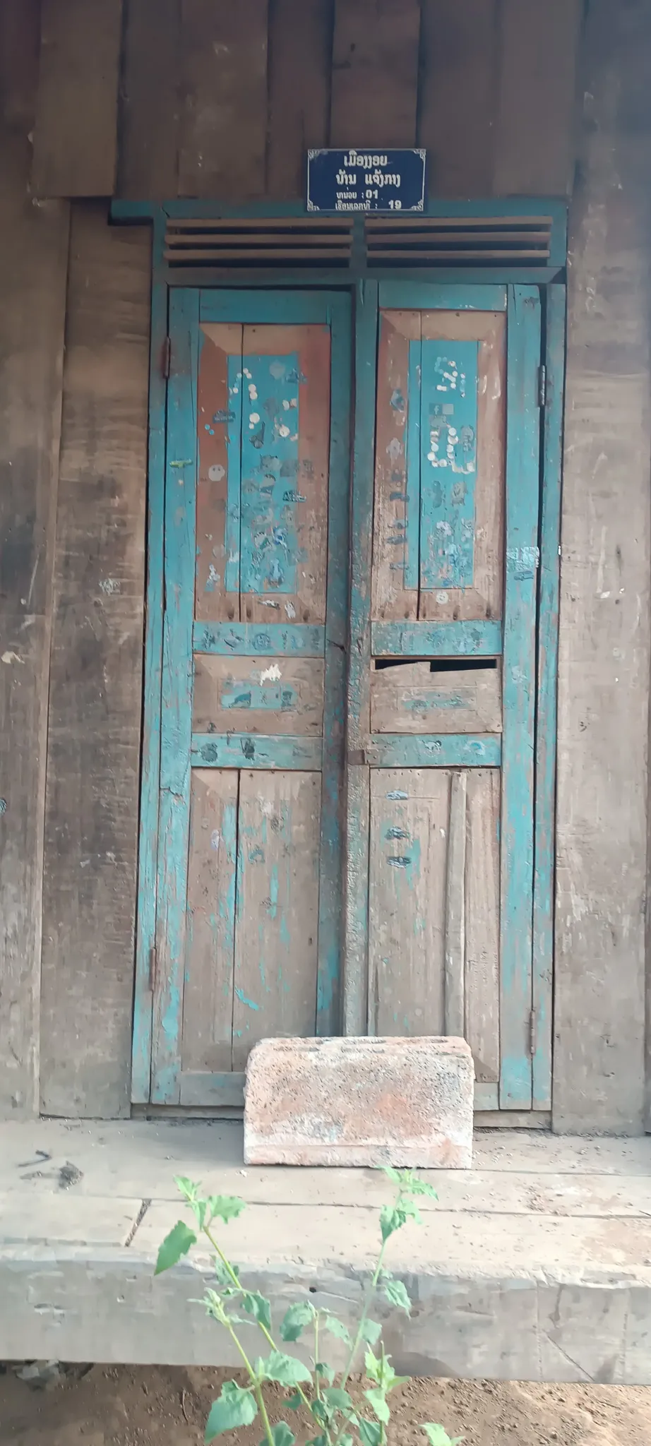 Weathered turquoise wooden door with Lao script address plaque on an abandoned building at a settlement along the Nam Ou river