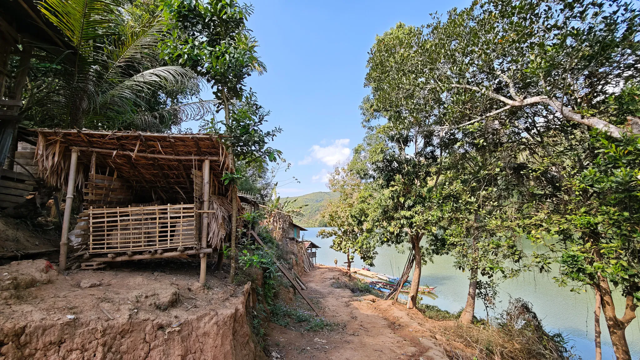 A raised bamboo hut beside a dirt path leading down to the Nam Ou river through a small Lao village