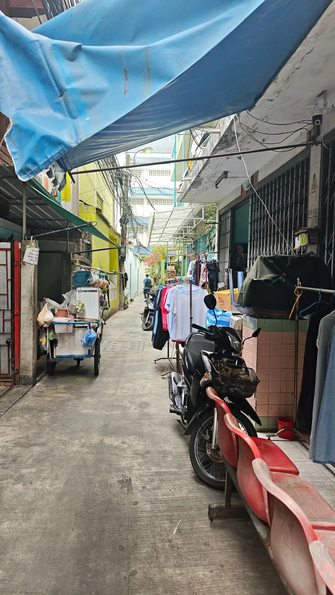 Bustling Bangkok back alley with a blue tarpaulin canopy, motorbikes, clothes racks and a street food cart