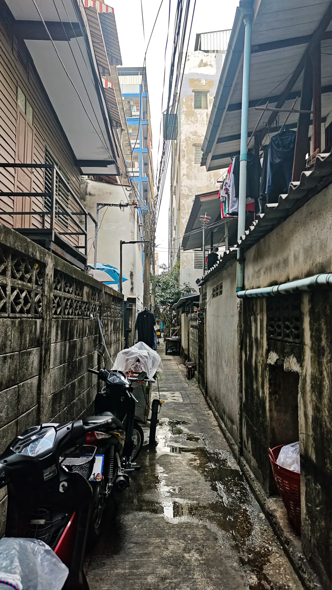 Narrow residential alleyway in Bangkok with motorbikes parked between concrete walls and laundry hanging overhead