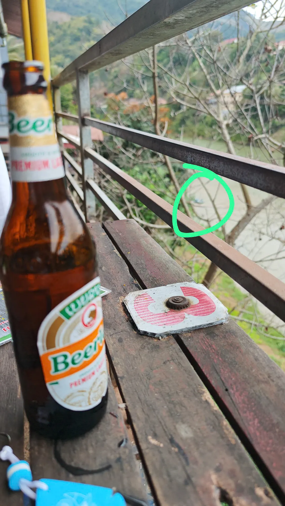 Two Beerlao bottles on a wooden balcony railing overlooking the Nam Ou river and surrounding hills in Muang Khua
