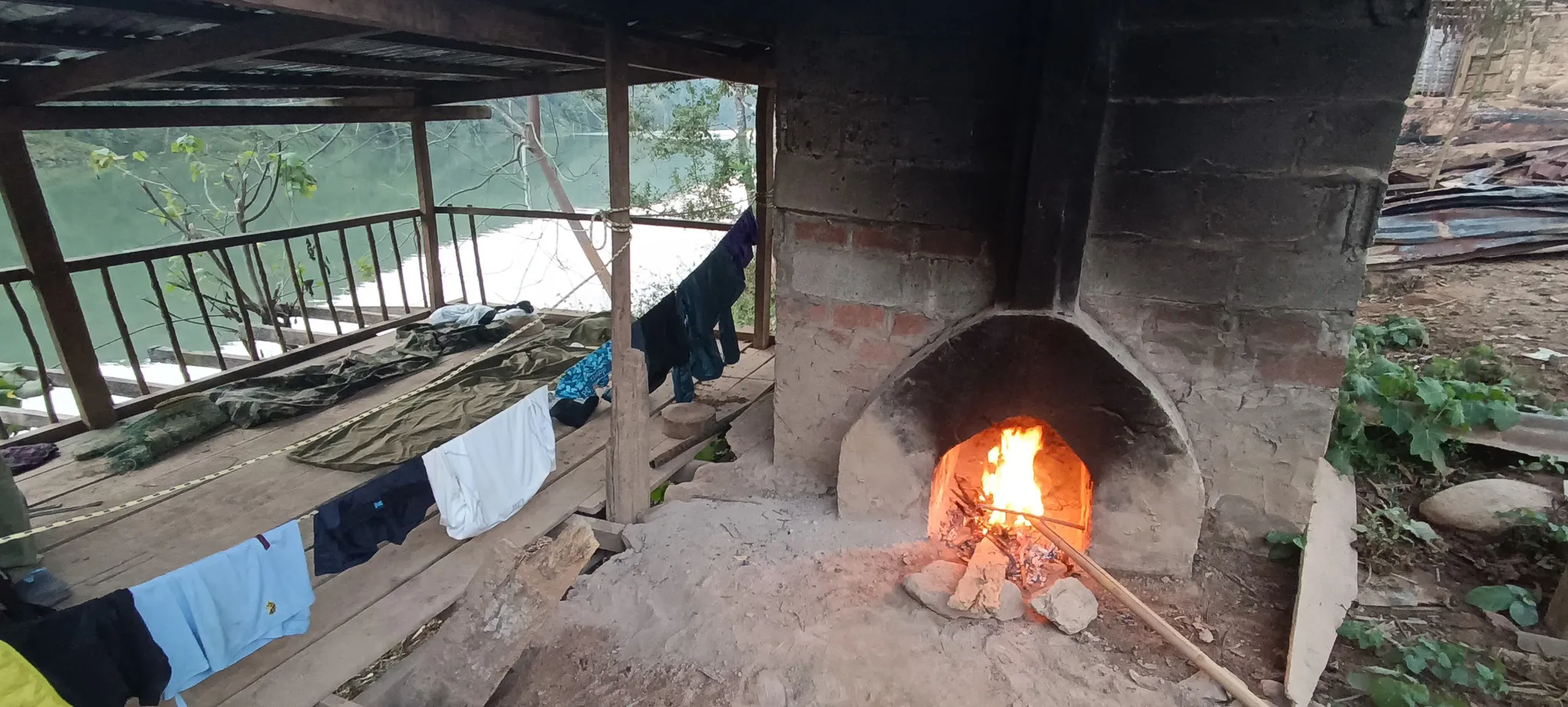 A fire burning inside a brick oven at the abandoned shelter camp with the Nam Ou river visible and clothes drying in the background