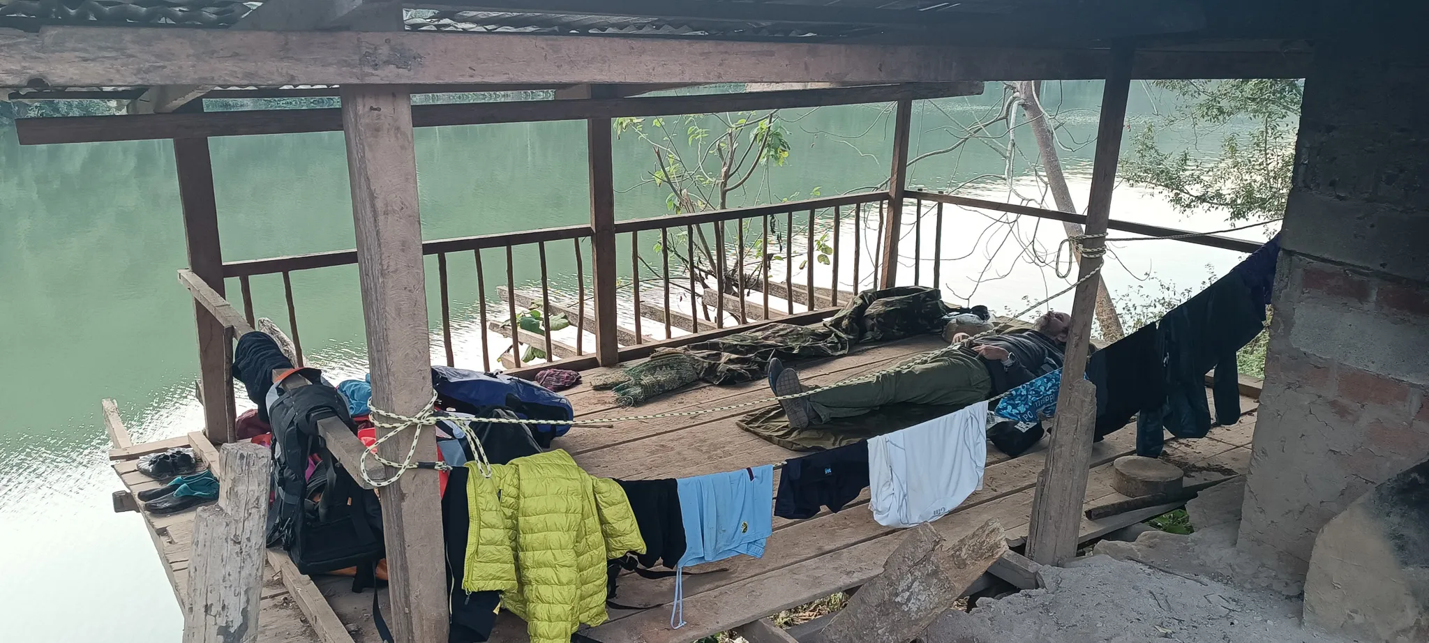 Bed rolls and sleeping bags laid out on the floor of a riverside shelter with the Nam Ou river visible through the railings