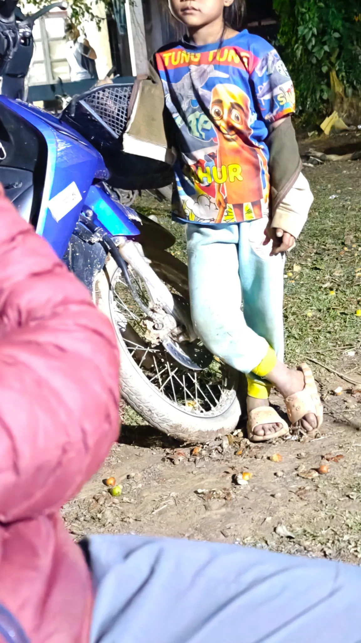 Child standing next to a blue motorbike at night in Ban Haddean village during the evening petanque gathering