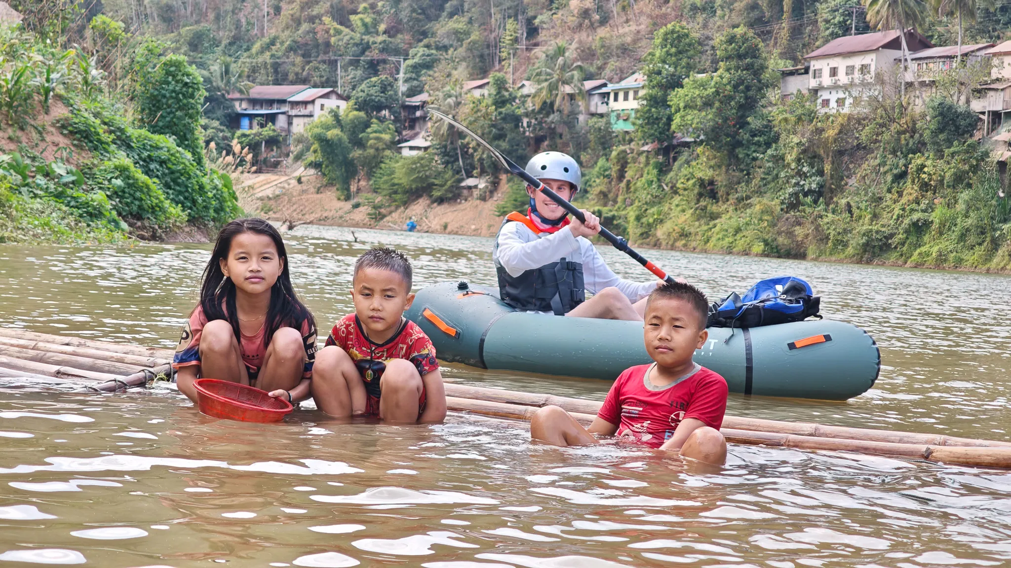 Three local children sitting in the Nam Ou river on a bamboo raft while a packrafter paddles past in an Itiwit raft near Muang Khua