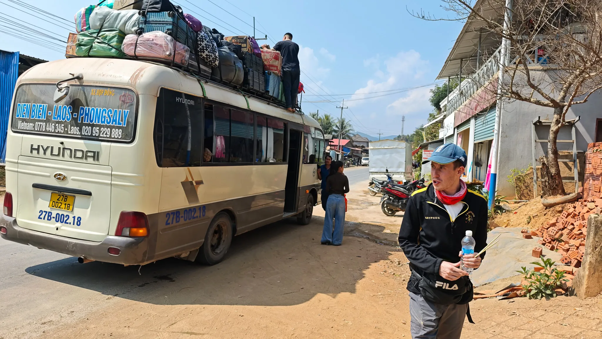 Rear view of the Dien Bien-Laos-Phongsaly bus showing the route sign and overloaded roof rack in a dusty Laos village