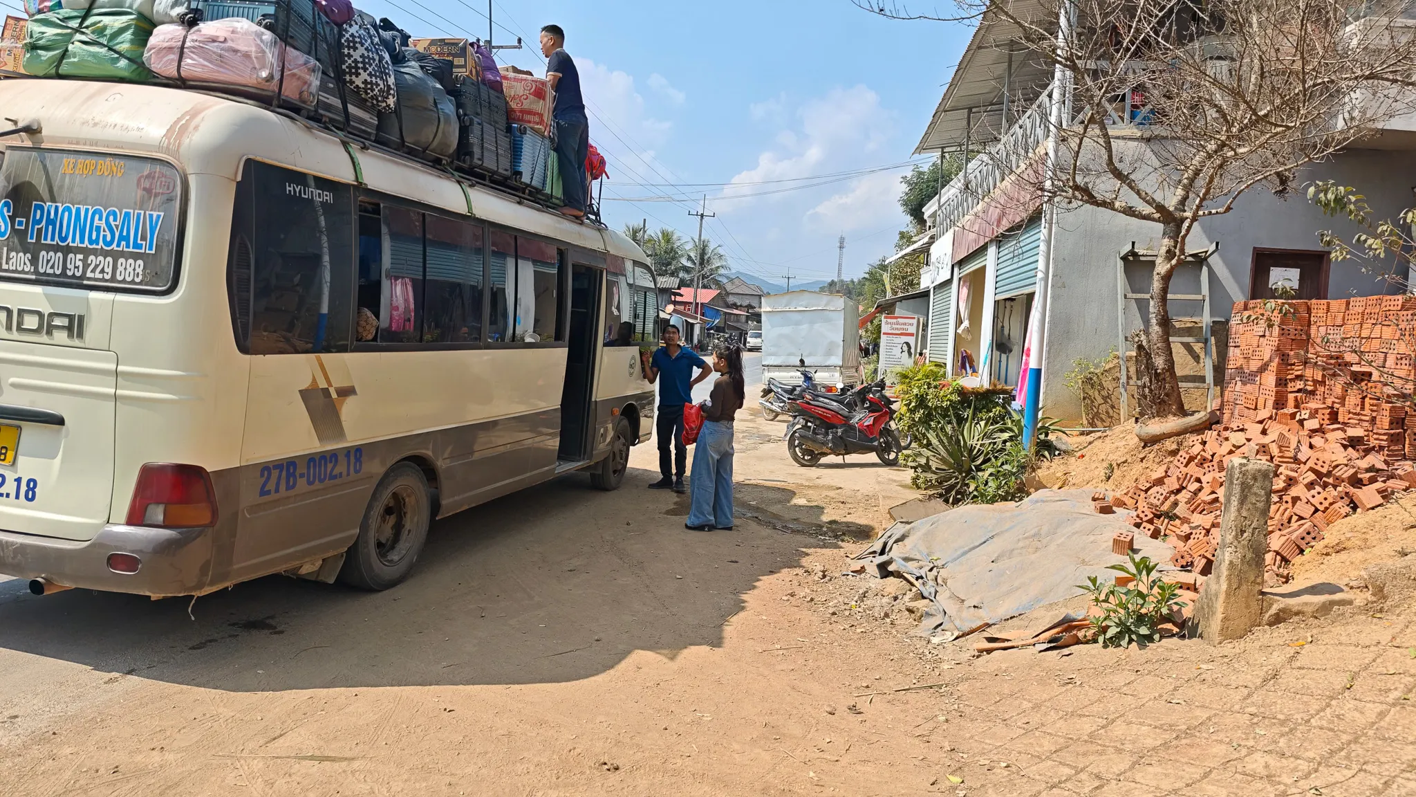 The Dien Bien to Phongsaly bus stopped in a Laos village with luggage piled on the roof and passengers stretching their legs