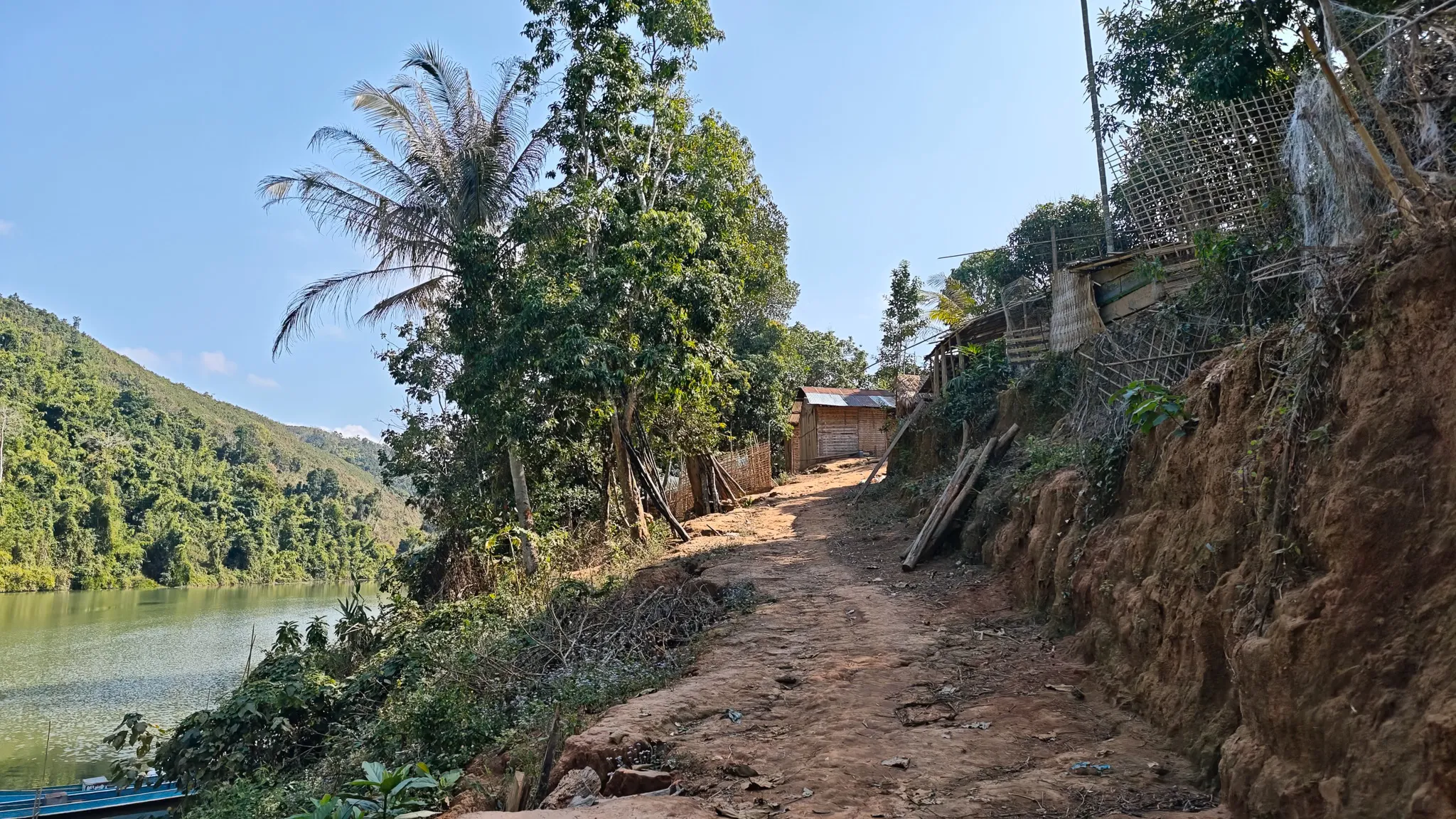 Narrow dirt path along the riverbank through a small village with wooden huts and palm trees beside the Nam Ou river