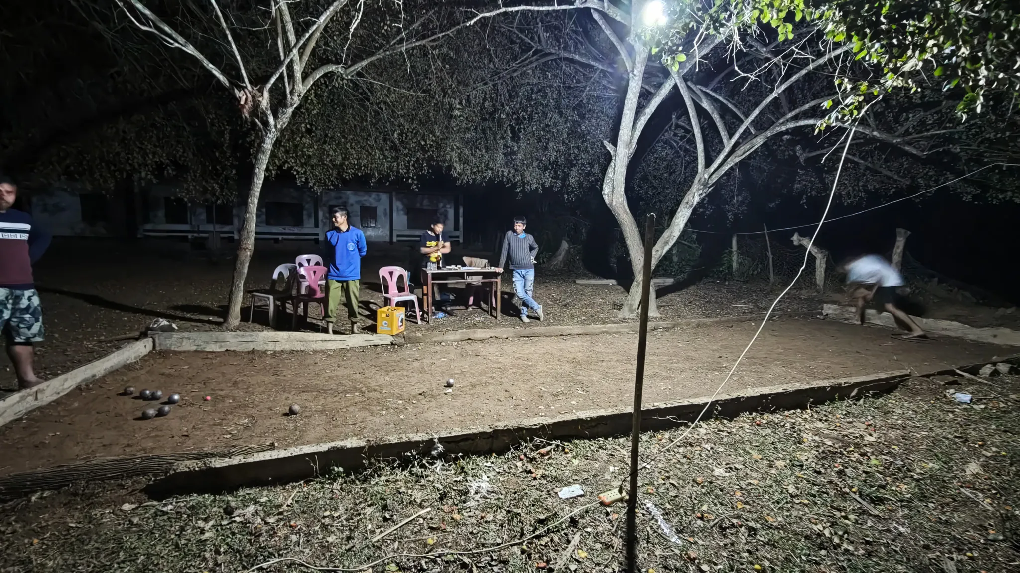 Evening petanque game under a single floodlight with locals gathered around tables and chairs in Ban Haddean village