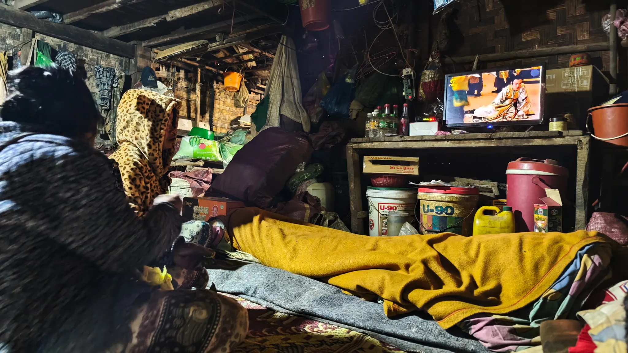 Villager wrapped in a blanket watching TV in a dimly lit wooden house at night in Ban Haddean on the Nam Ou