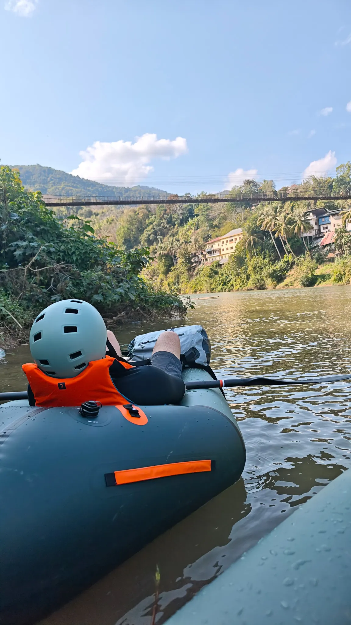 Point-of-view from a packraft showing feet up with the Muang Khua suspension bridge and riverside town in the background on the Nam Ou