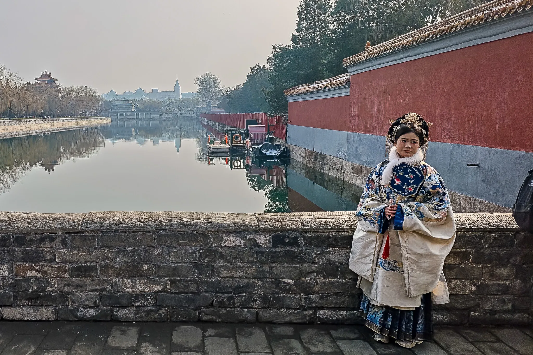 Woman in traditional Qing dynasty costume posing by the Forbidden City moat with red walls and a watchtower in the background