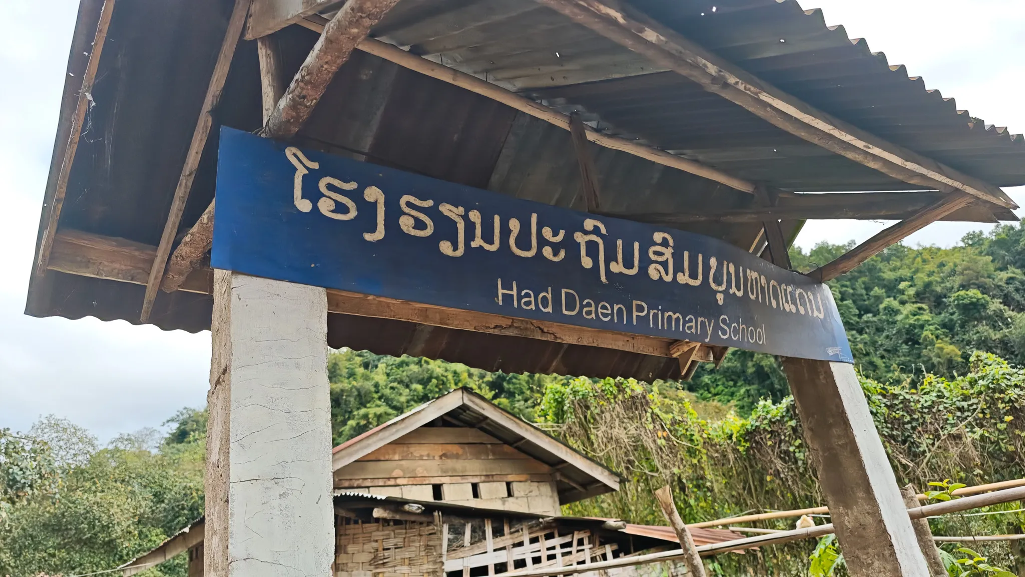 Had Daen Primary School entrance sign in Lao and English under a corrugated roof with forested hills behind, Phongsaly Province