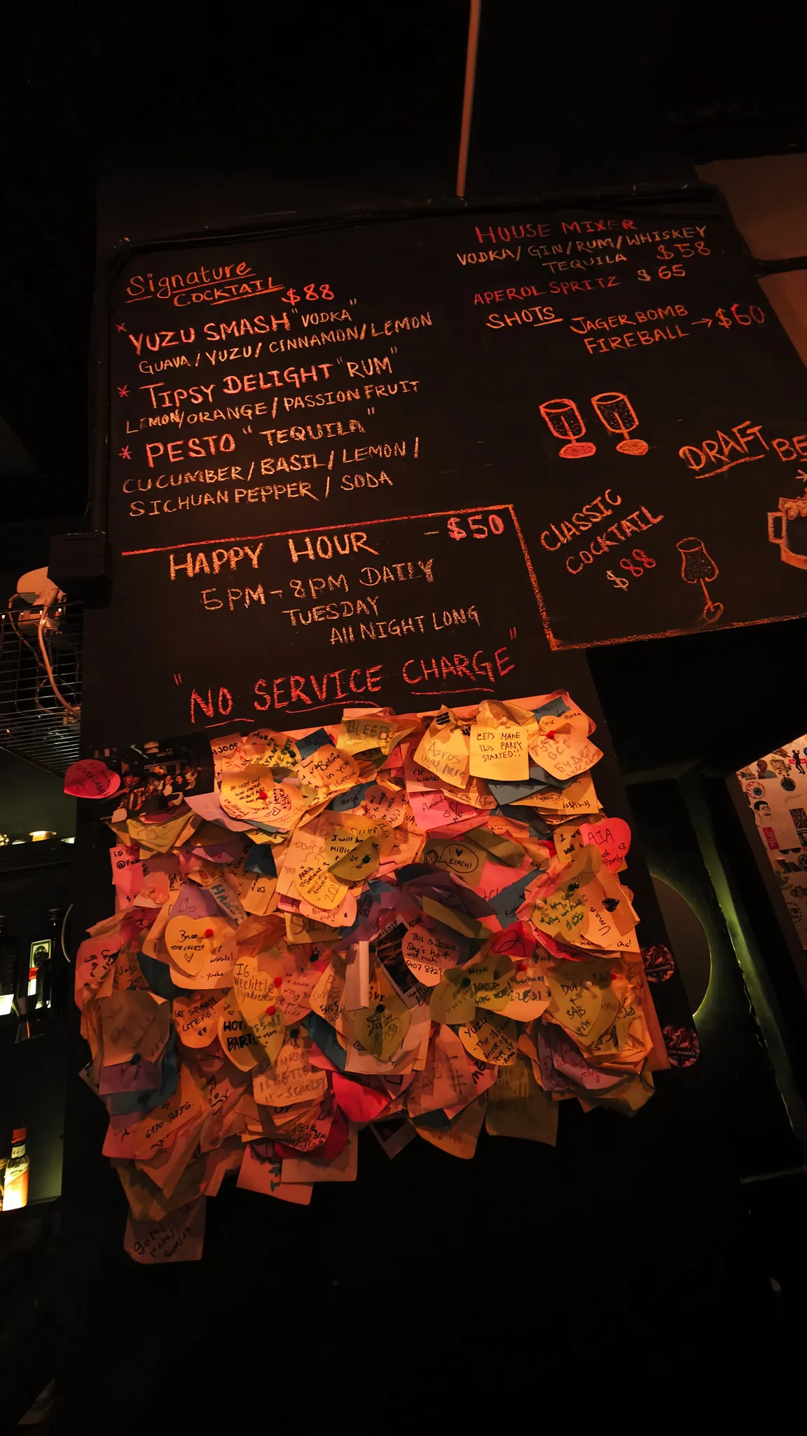 Chalkboard cocktail menu with signature drinks and happy hour prices, covered in heart-shaped sticky notes from visitors
