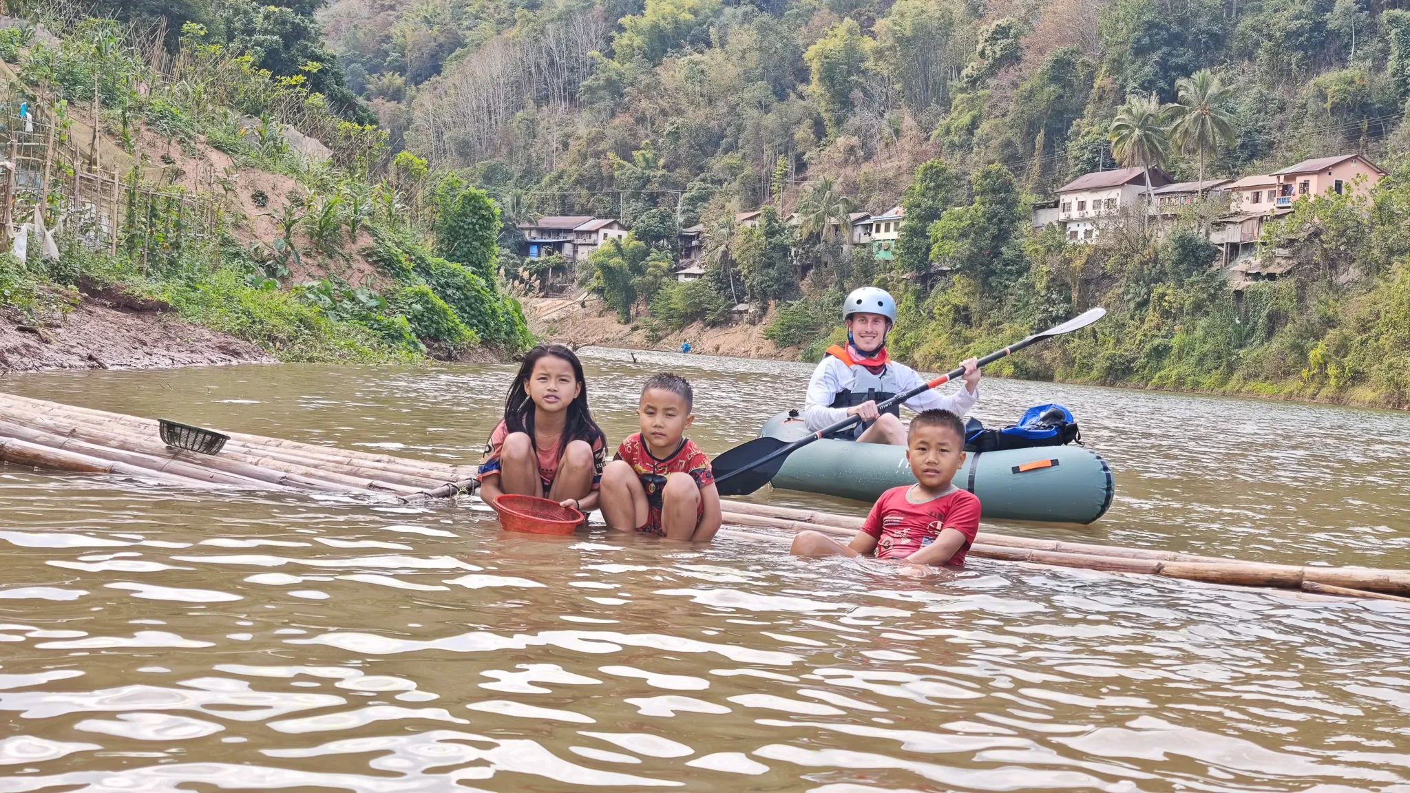 Lao children on a bamboo raft in the shallow water of the Nam Ou river with a packrafter approaching in the background at Muang Khua