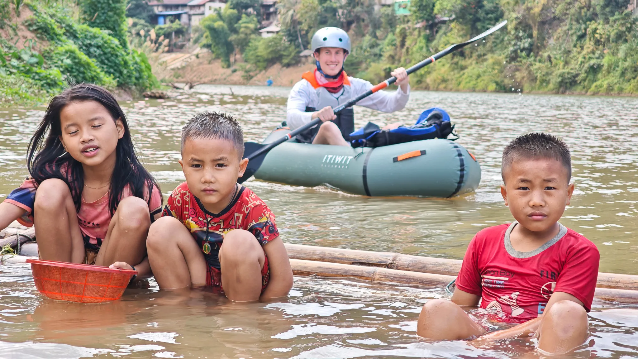 Close-up of three Lao children on a bamboo raft with an Itiwit packrafter smiling and raising his paddle on the Nam Ou river at Muang Khua