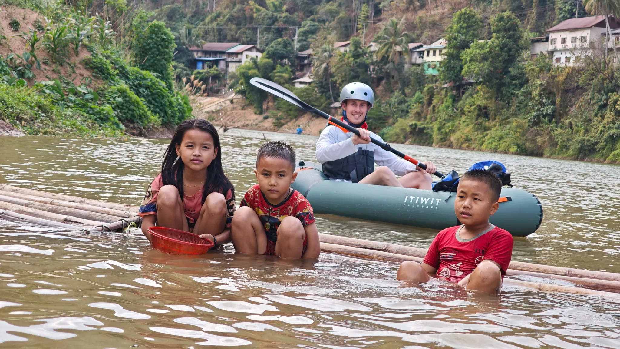 Three Lao children on a bamboo raft with a smiling Itiwit packrafter behind them on the Nam Ou river at Muang Khua