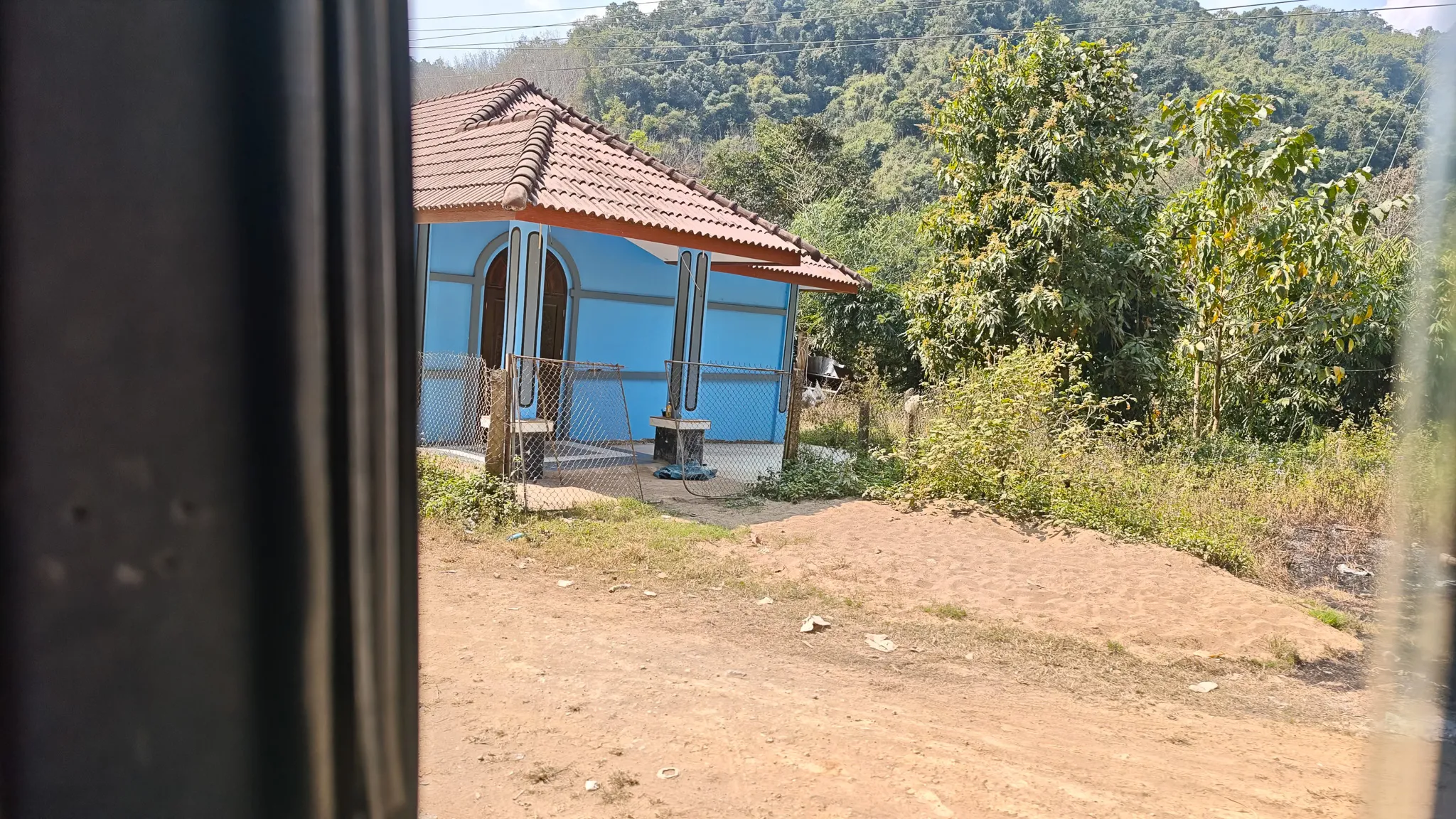 View from the bus window of a blue rural house with tile roof and forested hills in the northern Laos countryside