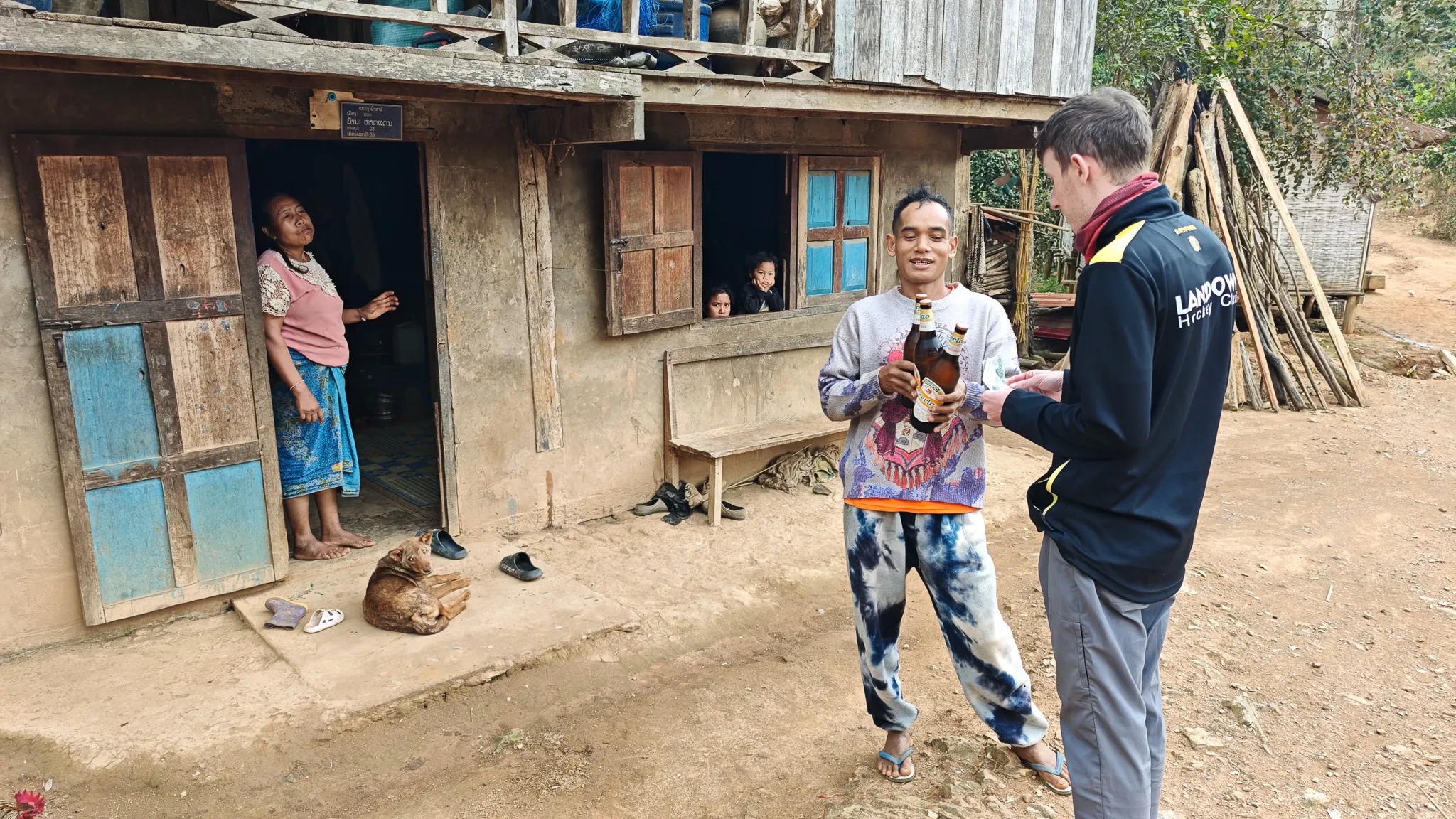 Local villager offering bottles of Beer Lao to a traveller outside a traditional wooden house in Ban Haddean, with a woman laughing in the doorway