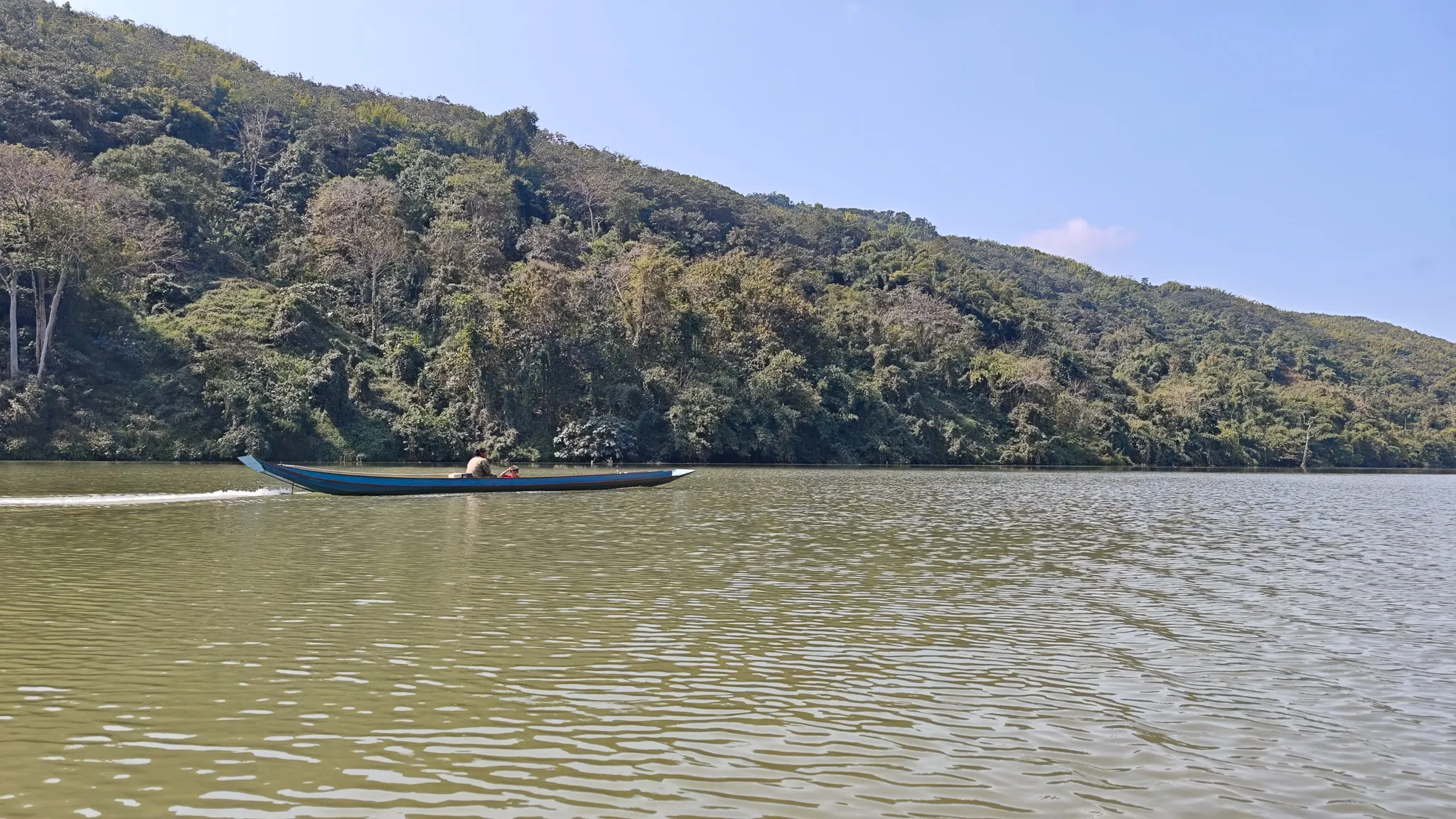 A traditional Lao longboat motoring past on the Nam Ou river with dense jungle hillside in the background