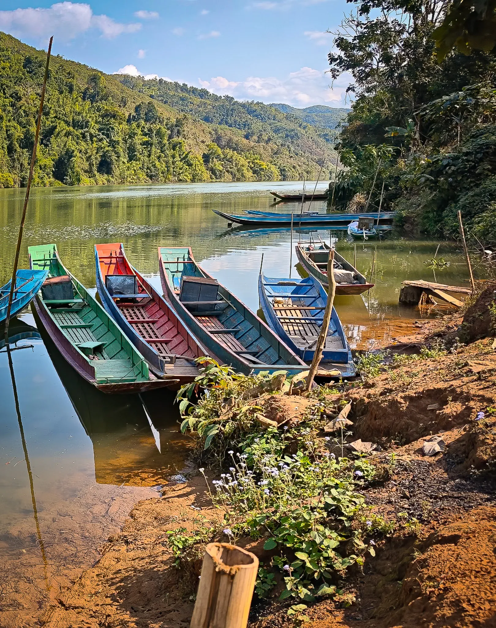 Row of colourful longboats and a bamboo raft moored along the Nam Ou river with forested hills reflected in calm water