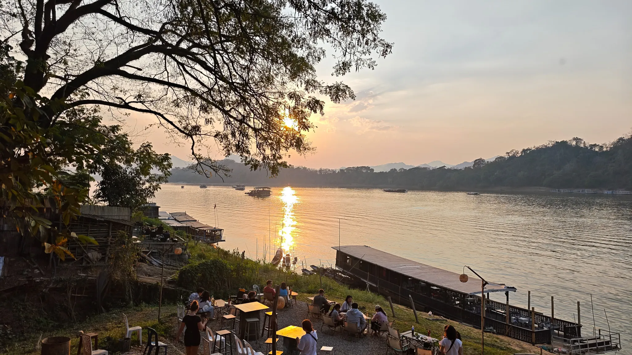 People relaxing at riverside cafes along the Mekong River during sunset in Luang Prabang with slow boats moored nearby
