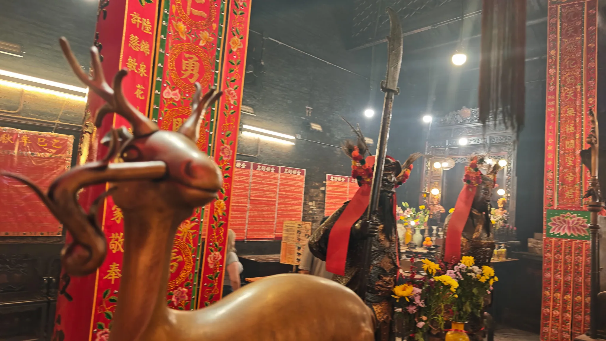 Bronze deer and warrior statue with a guandao blade surrounded by red banners and flower offerings inside Man Mo Temple