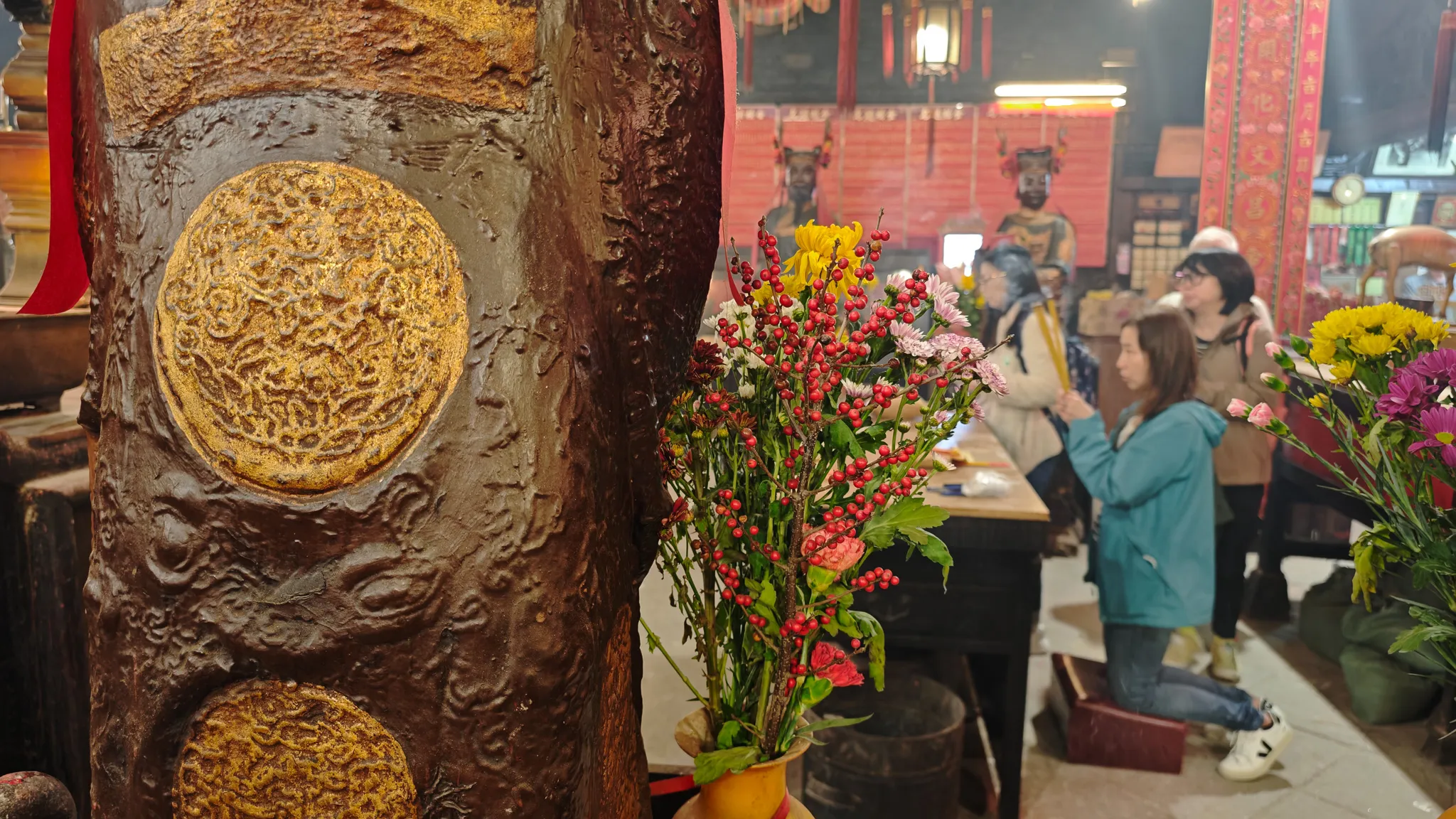 Close-up of an ornate bronze pillar with gold medallion inside Man Mo Temple, with worshippers and flower offerings in the background