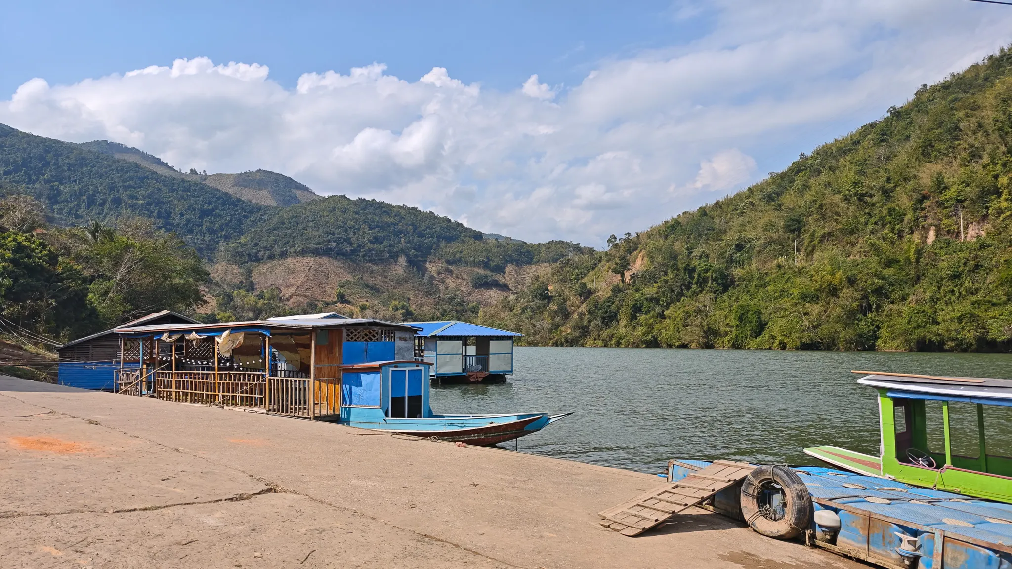 Boat landing at Muang Khua with a floating restaurant structure, colourful longboats, and forested mountains across the Nam Ou river