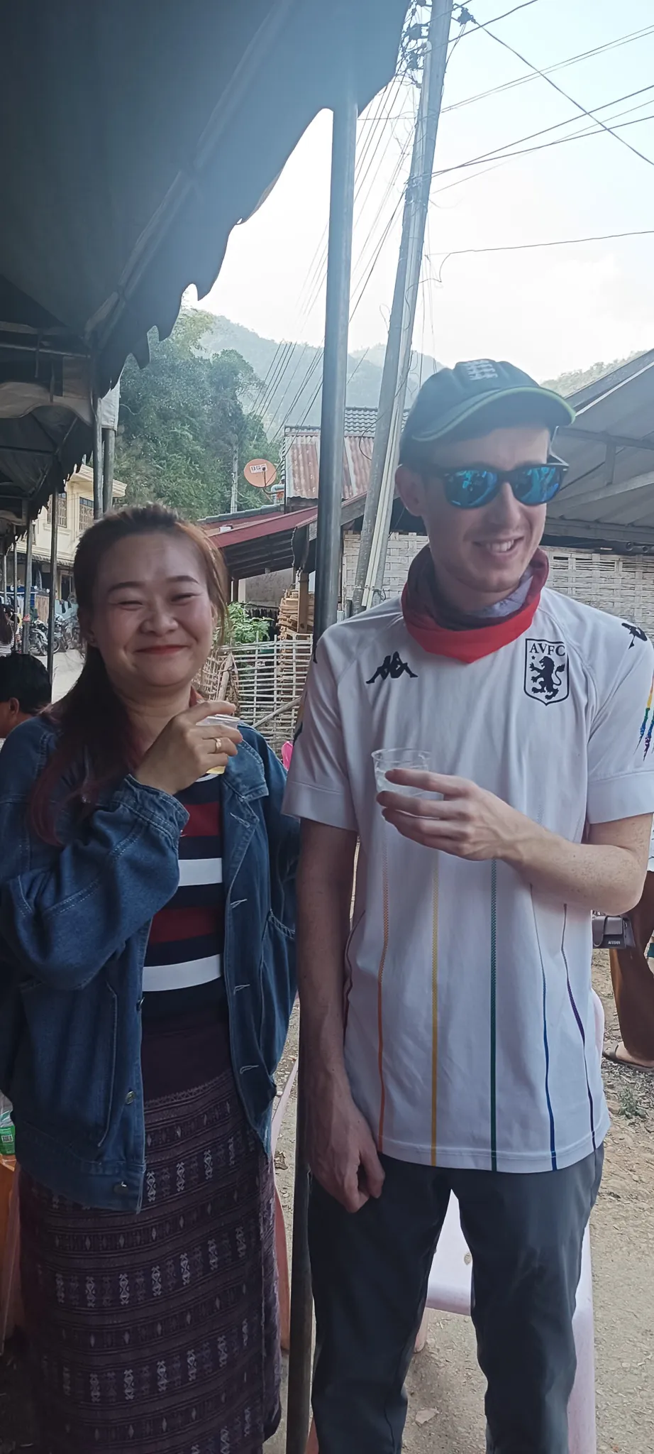 Traveller in an Aston Villa shirt drinking Lao whisky with a local woman in traditional sinh skirt at a community party in Muang Khua