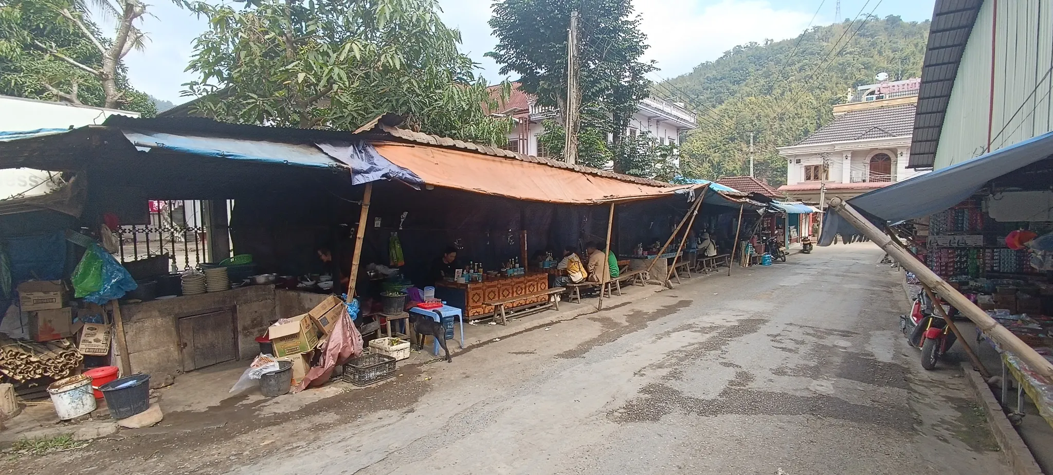 Muang Khua market street with covered food stalls, vendors cooking under tarpaulins, and mountains in the background