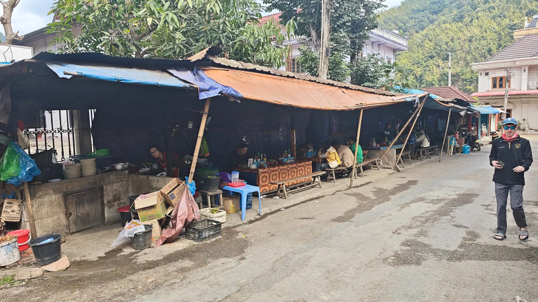 Traveller walking past the covered market stalls on the main street of Muang Khua with forested hills behind