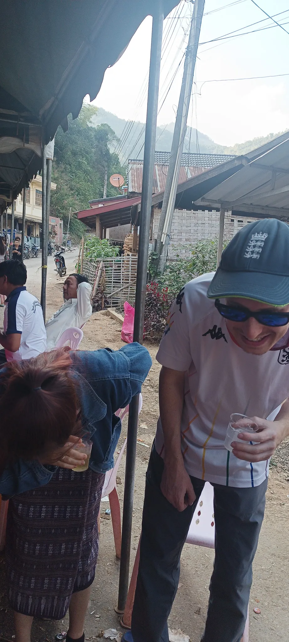 Traveller and local woman laughing while drinking Lao whisky at a community celebration under a tarpaulin in Muang Khua
