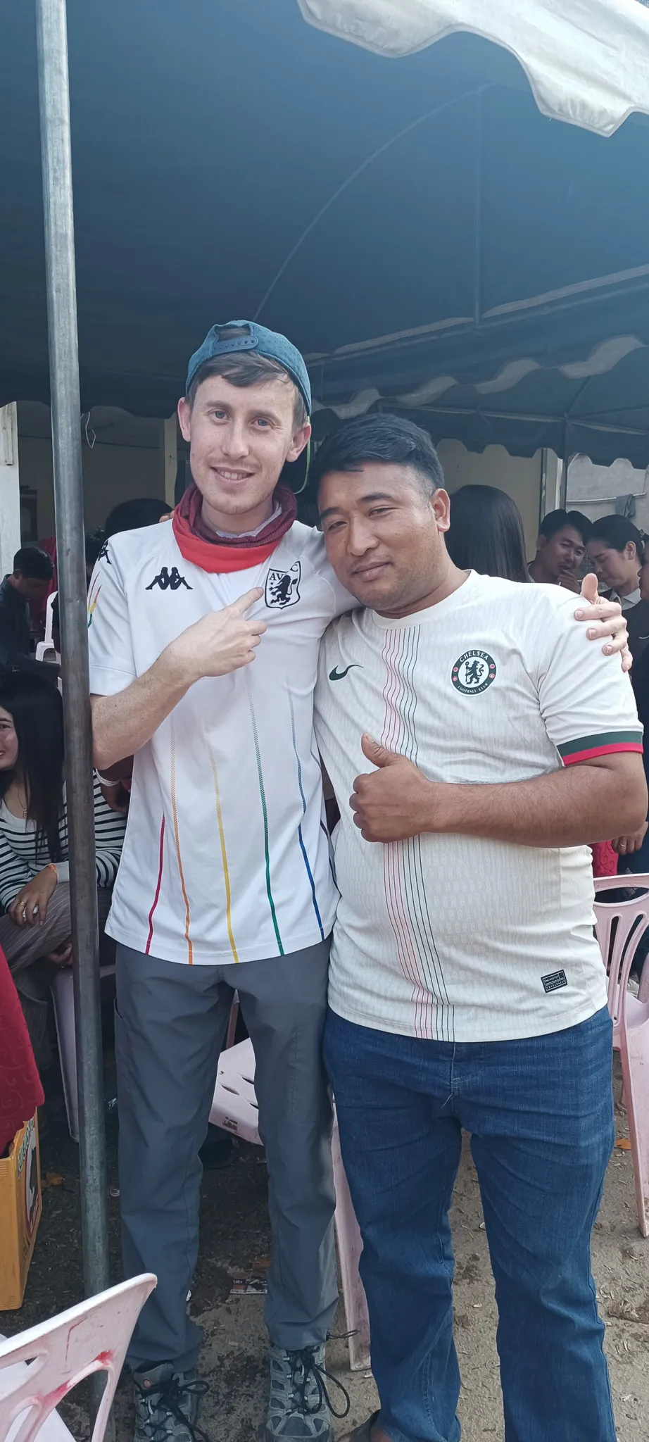 Traveller in Aston Villa shirt pointing at a local man in a Chelsea shirt, both smiling at the Muang Khua community party
