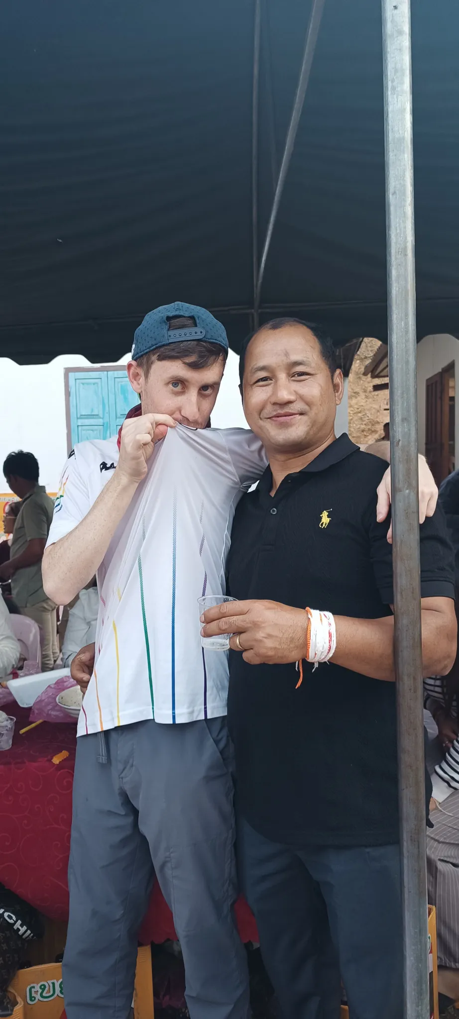 Traveller wiping his face with his shirt while posing with a local man holding Lao whisky at the Muang Khua community party