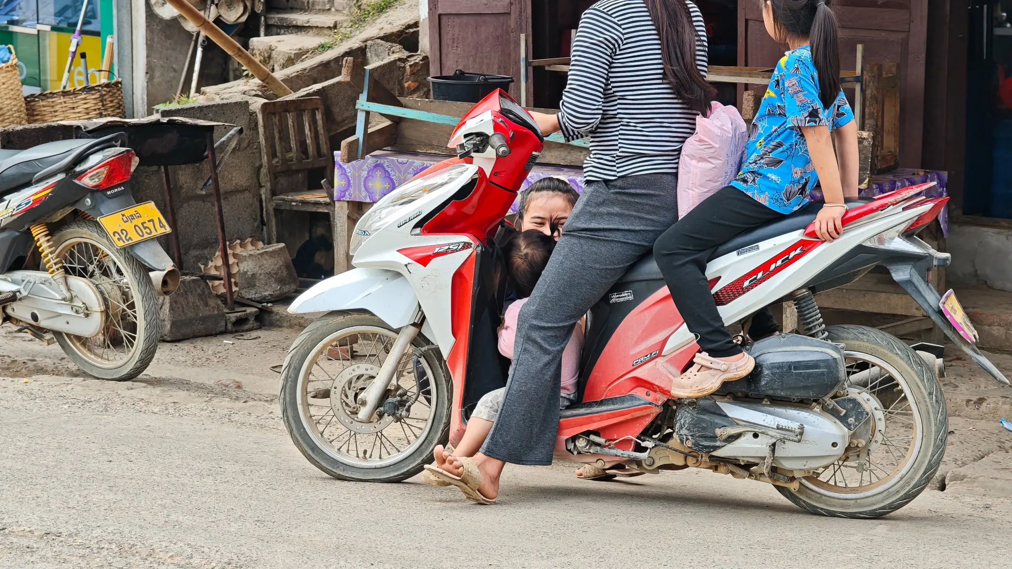 Three people on a motorbike on a street in Muang Khua with parked scooters and local shops in the background