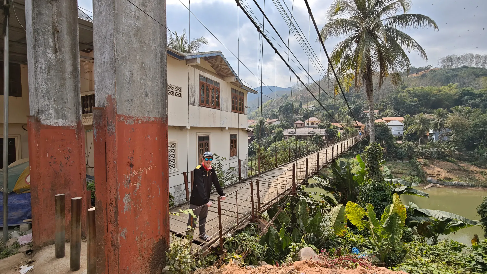 Traveller standing at the entrance of the rusty suspension bridge crossing the Nam Ou river in Muang Khua with palm trees and town behind