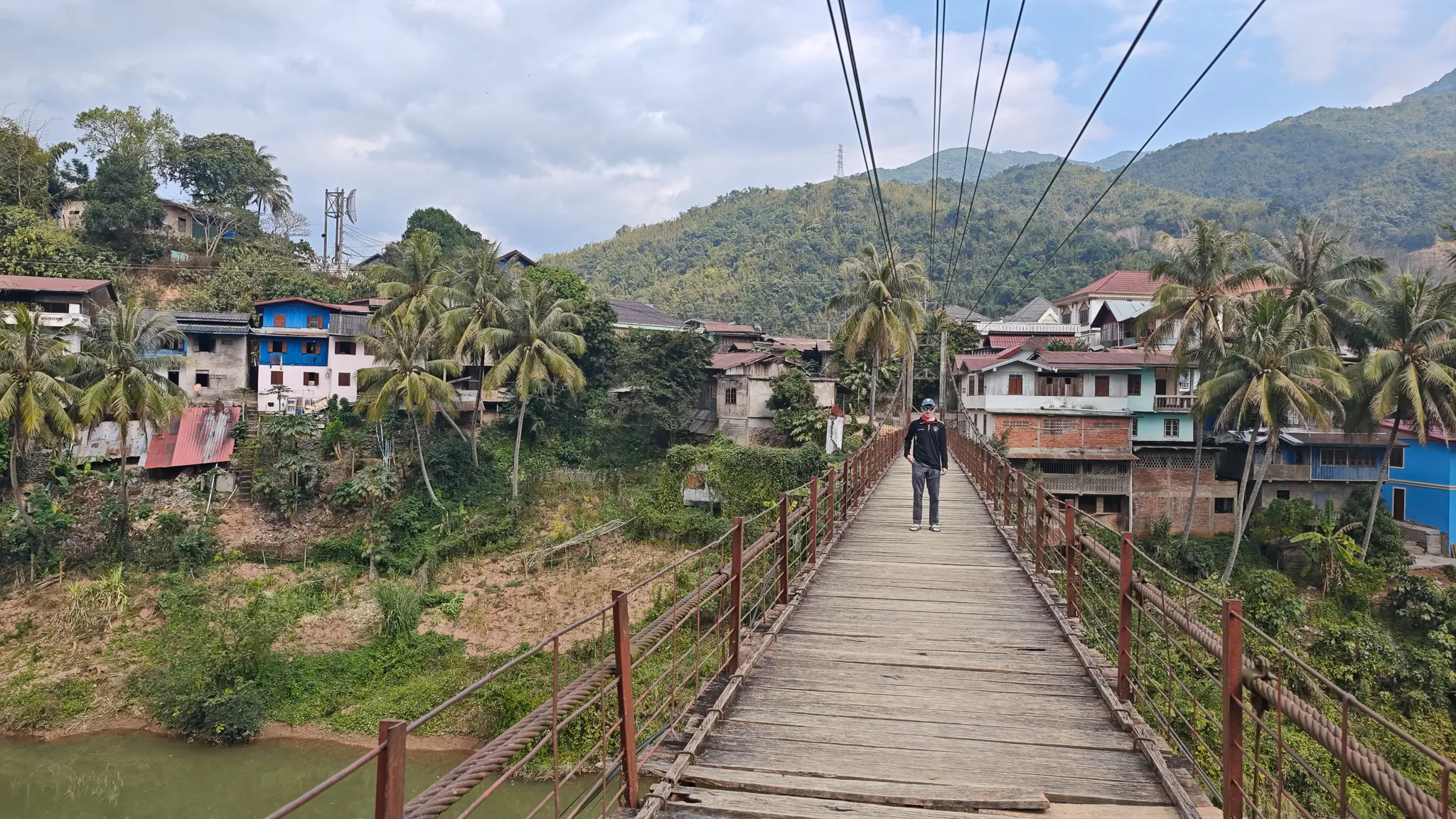 Traveller walking across the wooden-planked suspension bridge over the Nam Ou river in Muang Khua with town buildings ahead