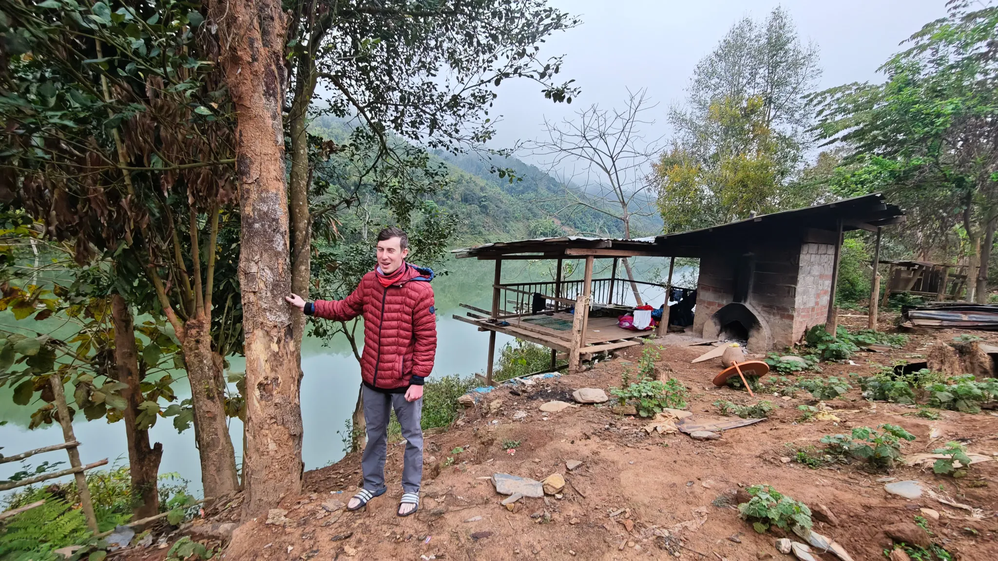 Packrafter in a red jacket standing beside a tree overlooking the Nam Ou river and a village shelter
