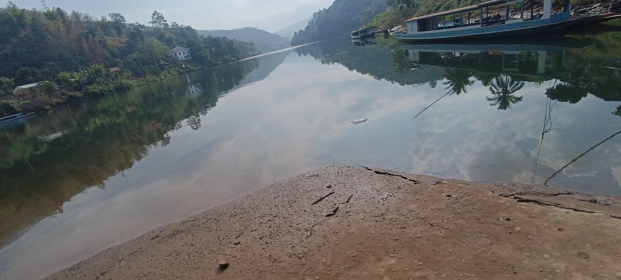 Wide view of the still Nam Ou river at Muang Khua with boats moored on the right bank and tree-covered hills on both sides