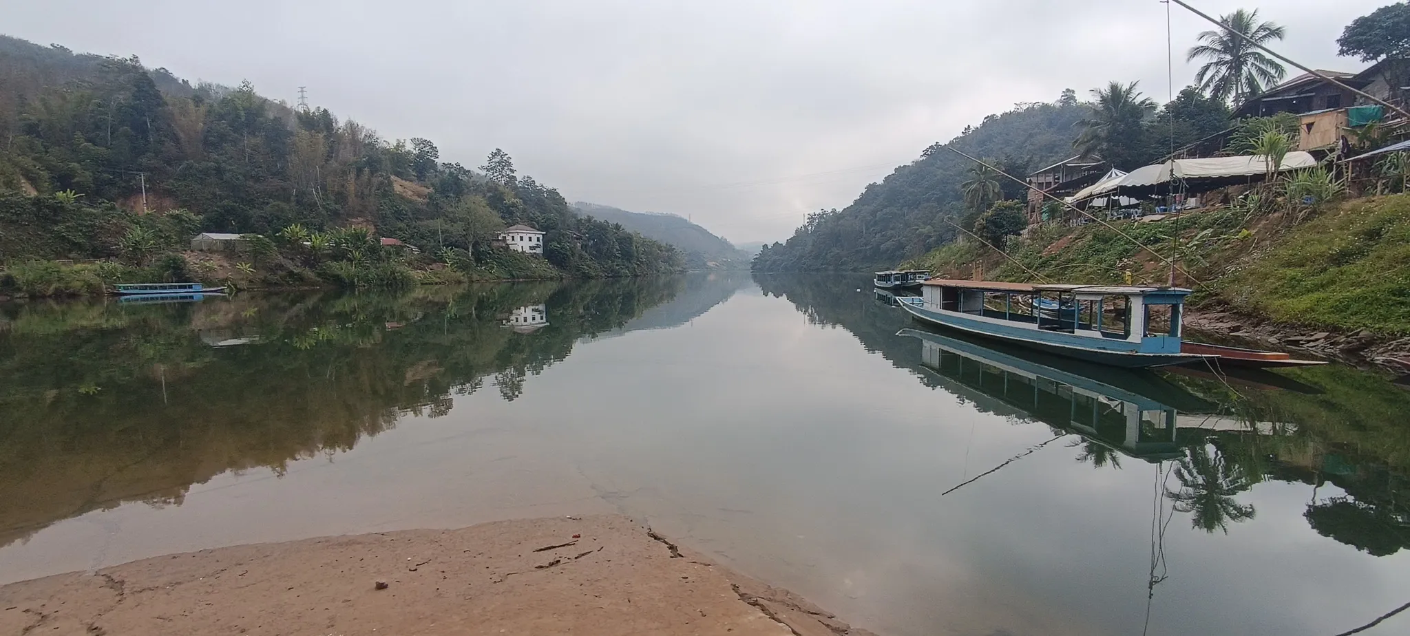 Calm morning view of the Nam Ou river at Muang Khua with longboats moored along the bank and misty forested hills