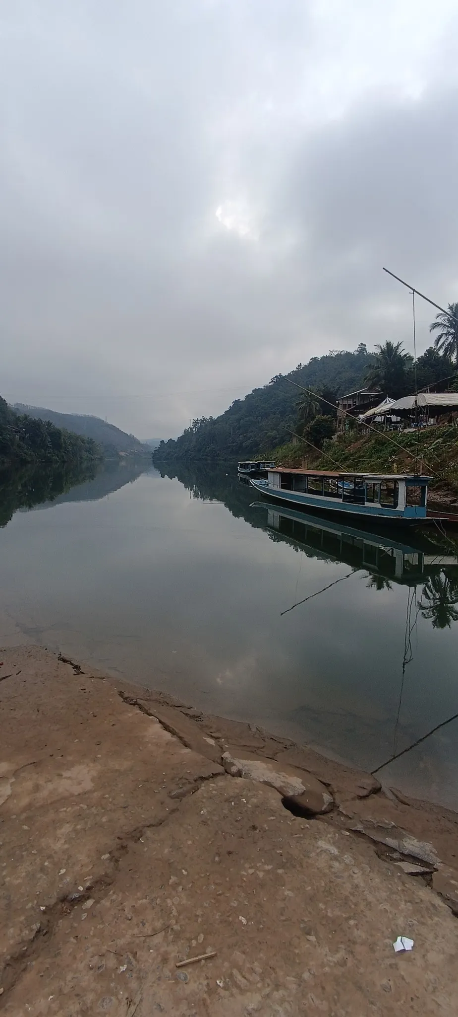 Misty morning on the Nam Ou river at Muang Khua with a traditional Lao longboat moored by the rocky riverbank