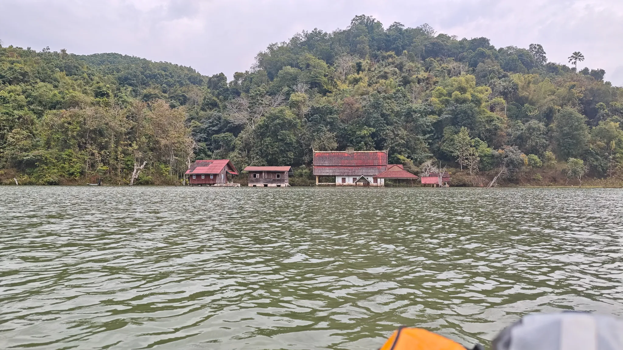 Red-roofed buildings nestled in dense jungle on the bank of the Nam Ou river, seen from an approaching kayak