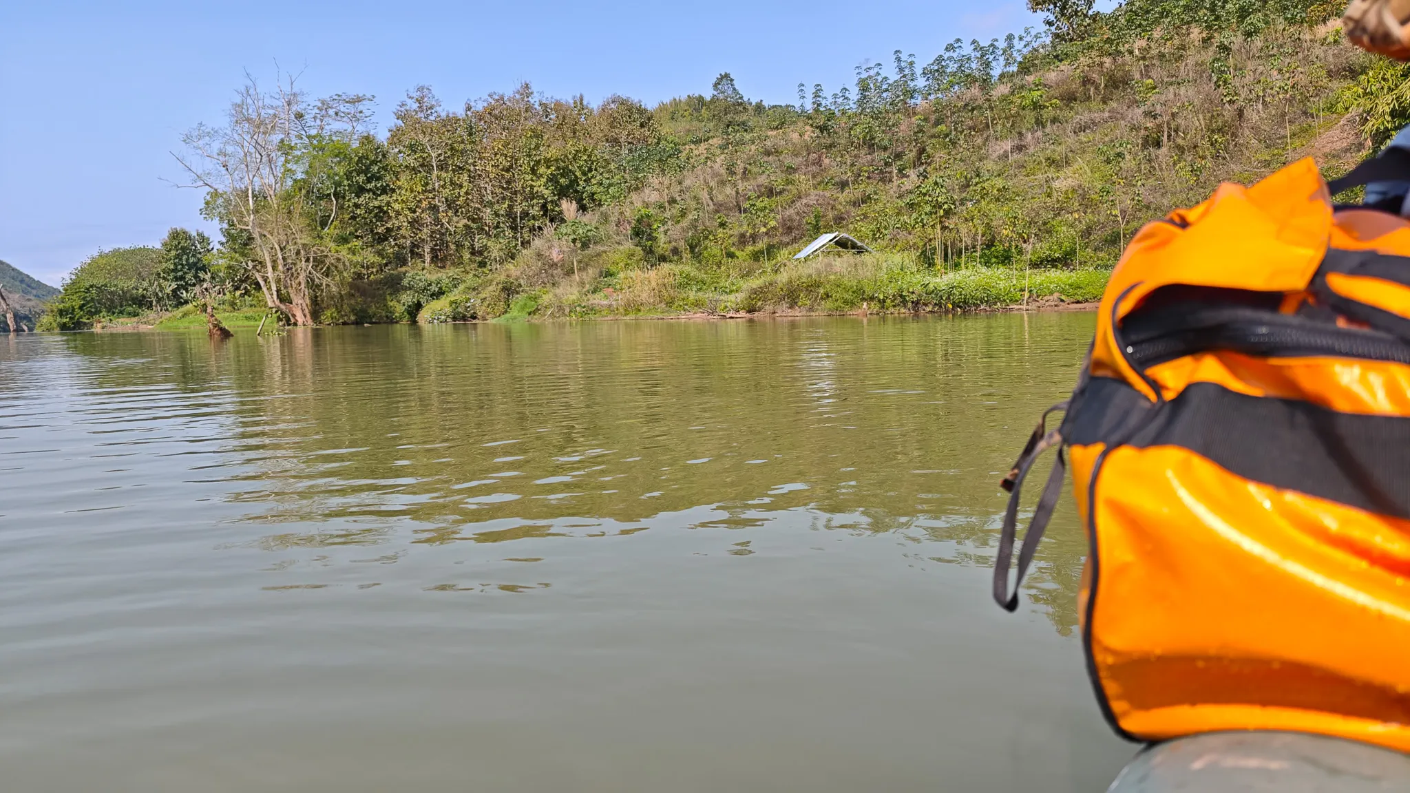 View from a packraft of a small riverside settlement with a tin roof building nestled among trees on the Nam Ou river hillside