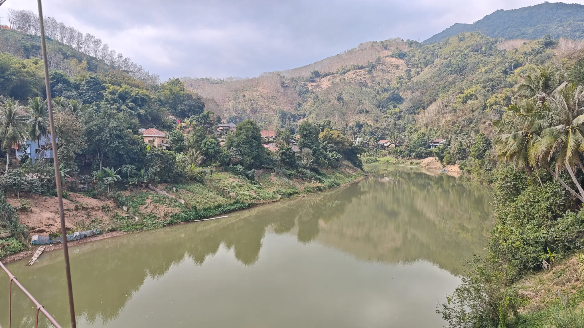 View of the Nam Ou river and surrounding village from the suspension bridge in Muang Khua, with forested mountains and palm trees