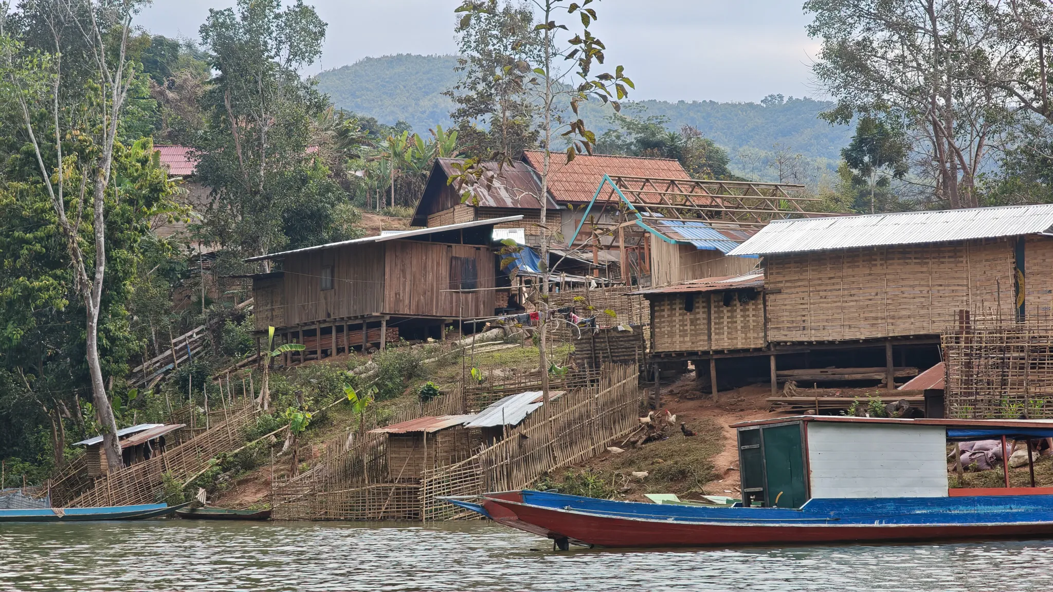 Hillside village with bamboo stilt houses, scaffolding and a blue longboat along the Nam Ou river bank