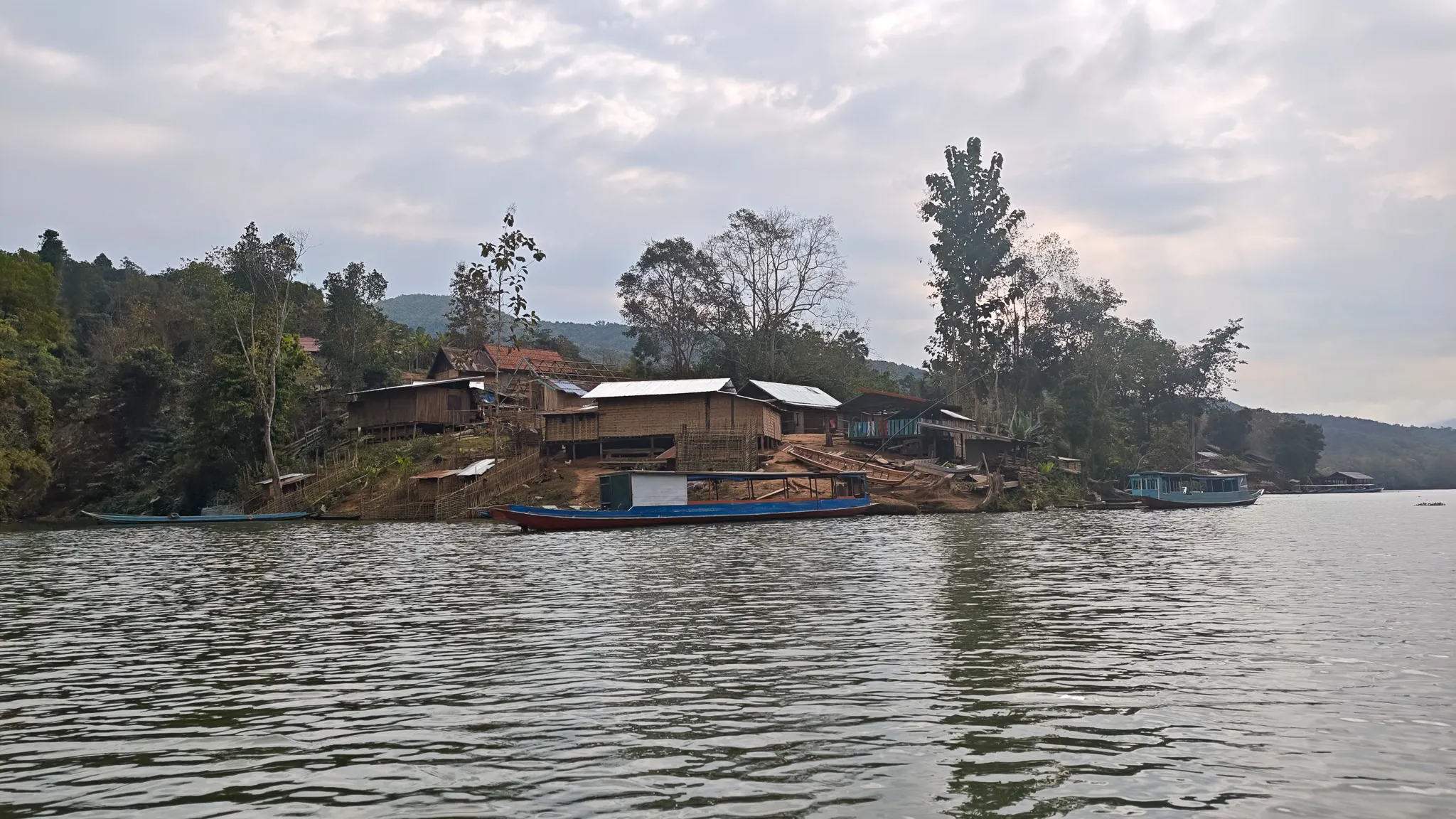 Riverside village with stilt houses and a blue longboat seen from the water while kayaking the Nam Ou