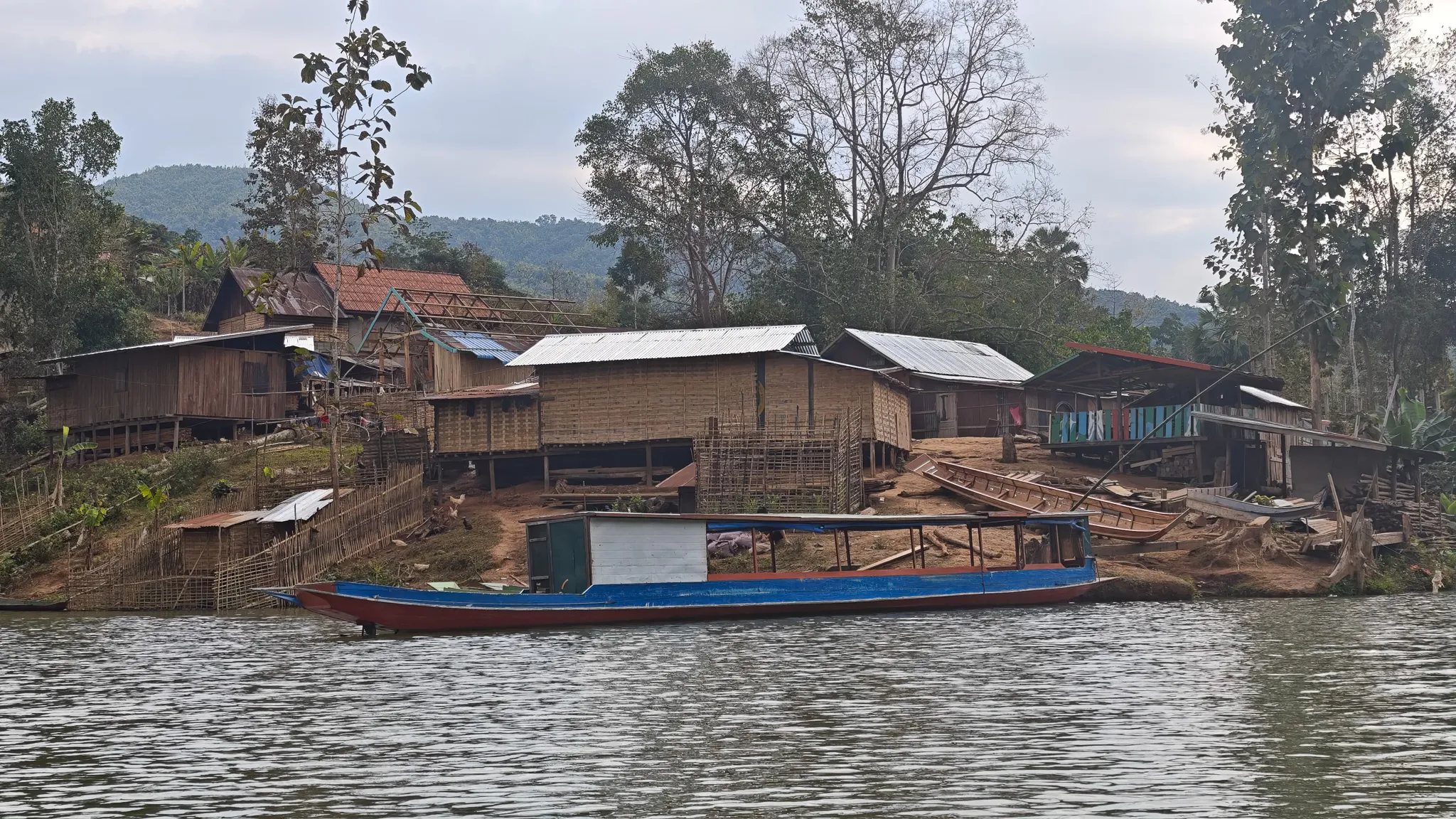 Closer view of bamboo and wooden stilt houses with a blue longboat moored at a Nam Ou river village