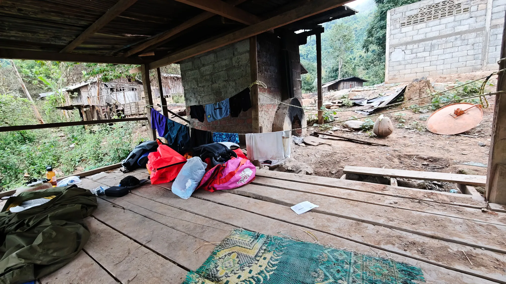 Camping gear spread out under a village shelter beside the Nam Ou river, with wooden houses in the background